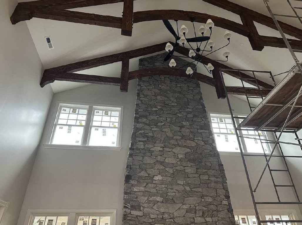 Vaulted great room with timber beams, stone fireplace and chandelier during construction